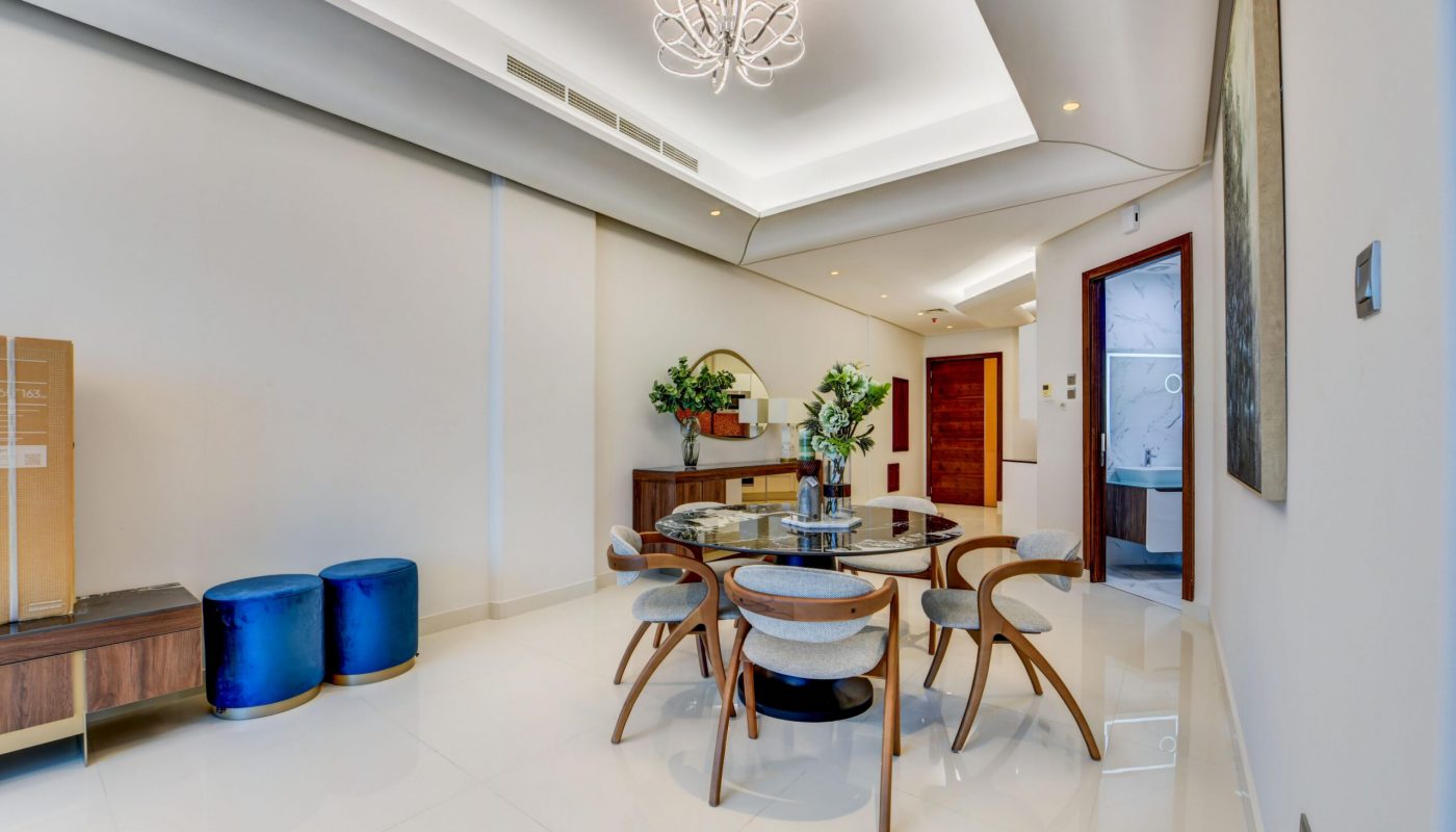 A modern dining room with a round glass table, six chairs, decorative vases, blue stools, wooden accents, and a chandelier, with an open door leading to another room.