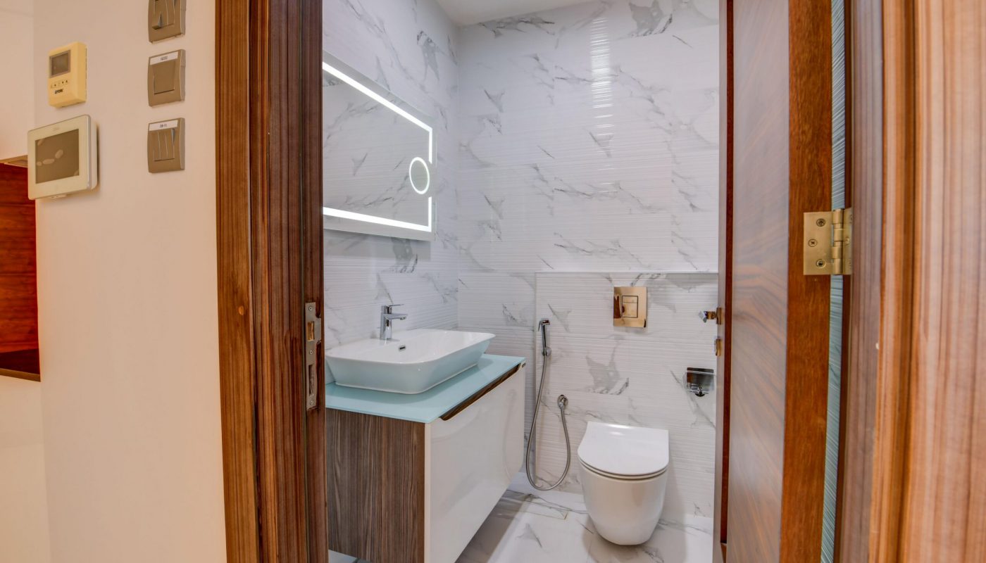 Modern bathroom with white marble walls and floor, a wall-mounted sink with mirror, toilet, bidet spray, and wooden door partially open.