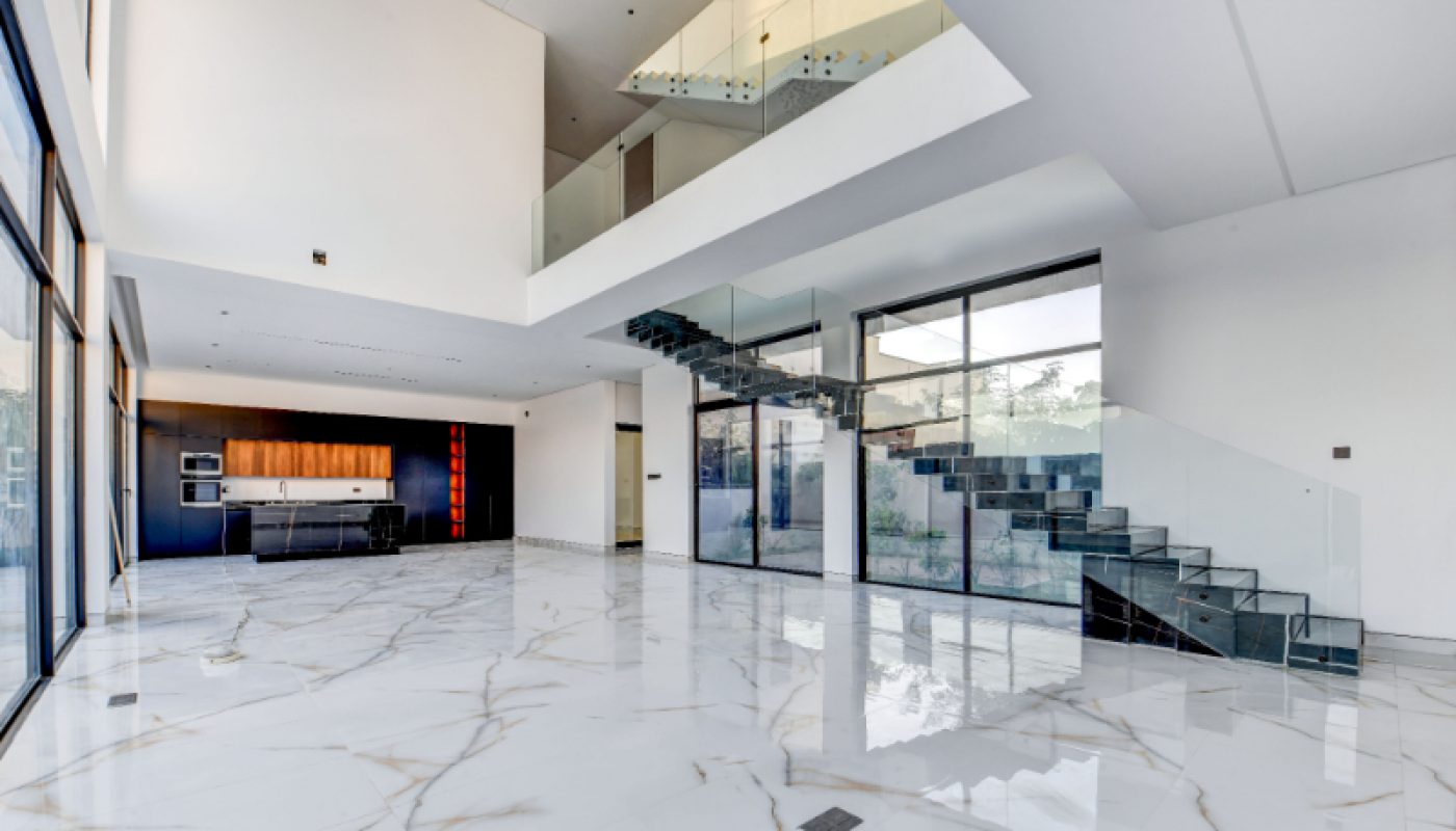 Spacious modern living area with large marble floors, glass staircase, floor-to-ceiling windows, and a view of the open kitchen in the background.