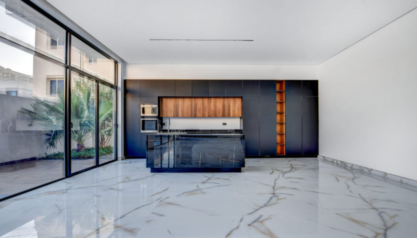 Modern kitchen with dark cabinets, built-in appliances, a central island, and large marble flooring; floor-to-ceiling windows reveal an outdoor patio with plants.