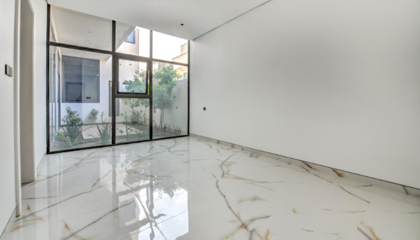Empty room with large floor-to-ceiling windows, white marble floor with gold veins, and white walls; garden area visible outside the windows.