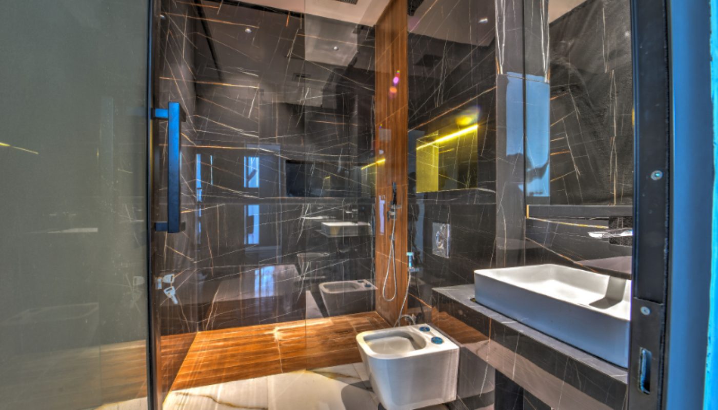 Modern bathroom with black marble walls, glass shower enclosure, wooden floor section, wall-mounted toilet, bidet, and rectangular sink with mirror and under-cabinet lighting.