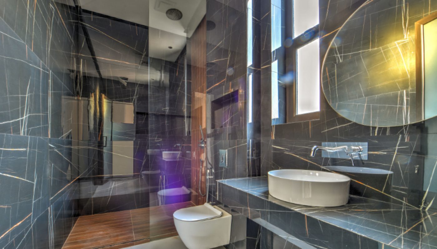 Modern bathroom with dark marble walls, wall-mounted toilet, round vessel sink, large round mirror, and walk-in shower with wooden floor. Natural light enters through tall windows.