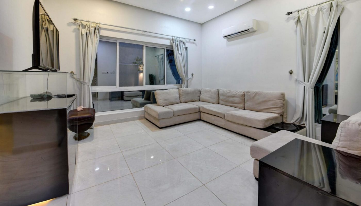 Modern living room with white tile flooring, cream sectional sofa, dark wood furniture, glass display cabinet, large windows, white curtains, and recessed ceiling lights.