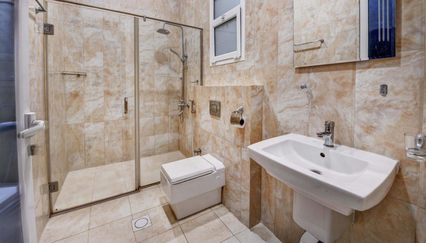 Modern bathroom with beige tiles, a wall-mounted sink, mirror, toilet, and a large glass-enclosed shower; ceiling features a patterned light panel.