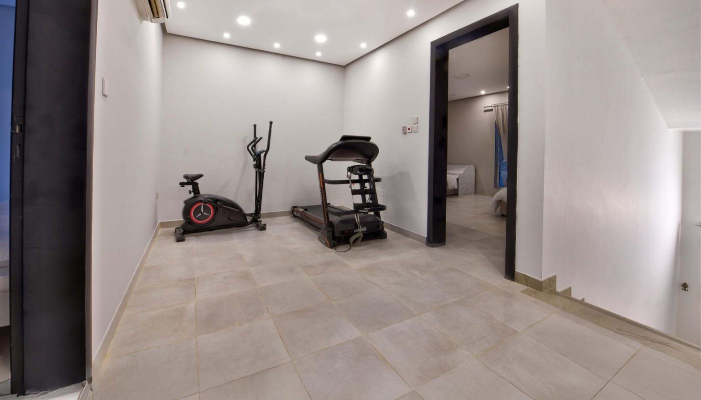 A small indoor gym room with an elliptical machine and a treadmill on tiled flooring, with white walls and recessed ceiling lights.