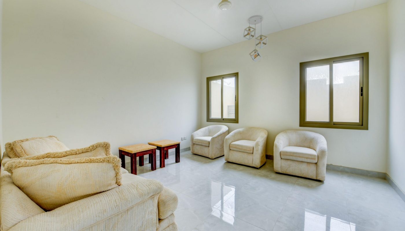 Minimalist living room with cream-colored armchairs and sofa, two small wooden tables, tiled floor, and two windows letting in natural light.