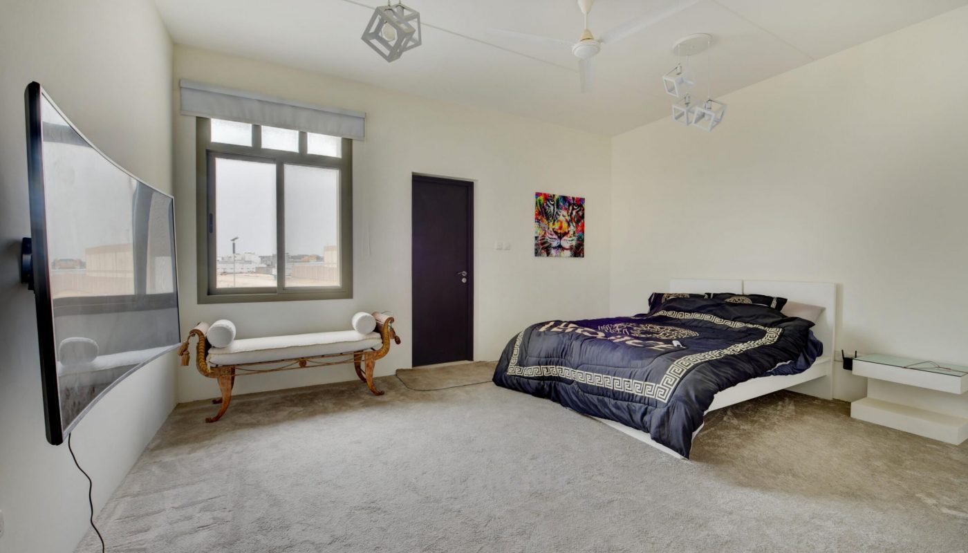 Modern bedroom with a large bed, a wall-mounted TV, a bench by the window, and minimalist decor. The room has light-colored walls and carpeted flooring.