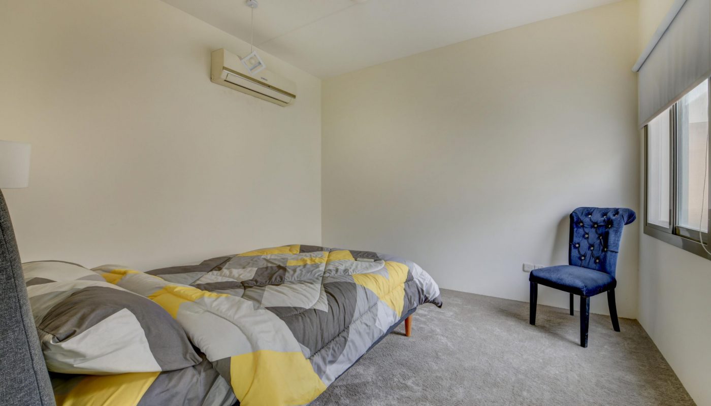 Minimalist bedroom with a bed covered in a geometric-patterned comforter, a blue chair by the window, and an air conditioner mounted on the wall.