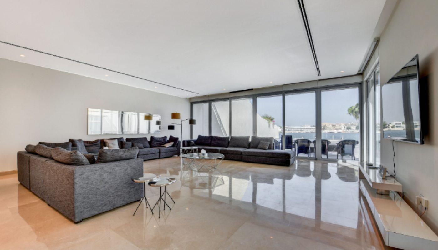 Spacious modern living room with large gray sectional sofas, marble flooring, wall-mounted TV, and floor-to-ceiling windows overlooking a balcony with waterfront views.