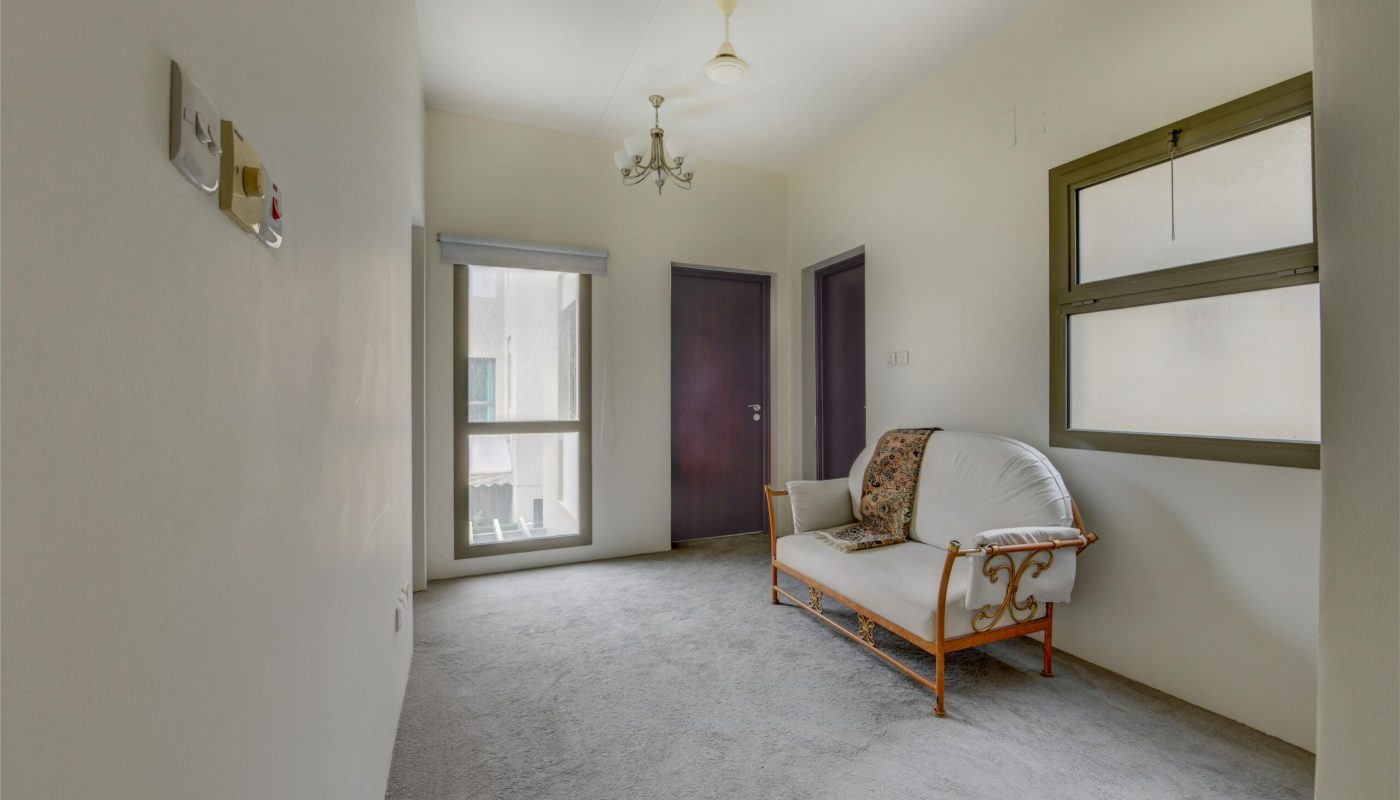 A small, carpeted room with a window, two closed doors, a light fixture, and a white metal sofa with cushions against the wall.
