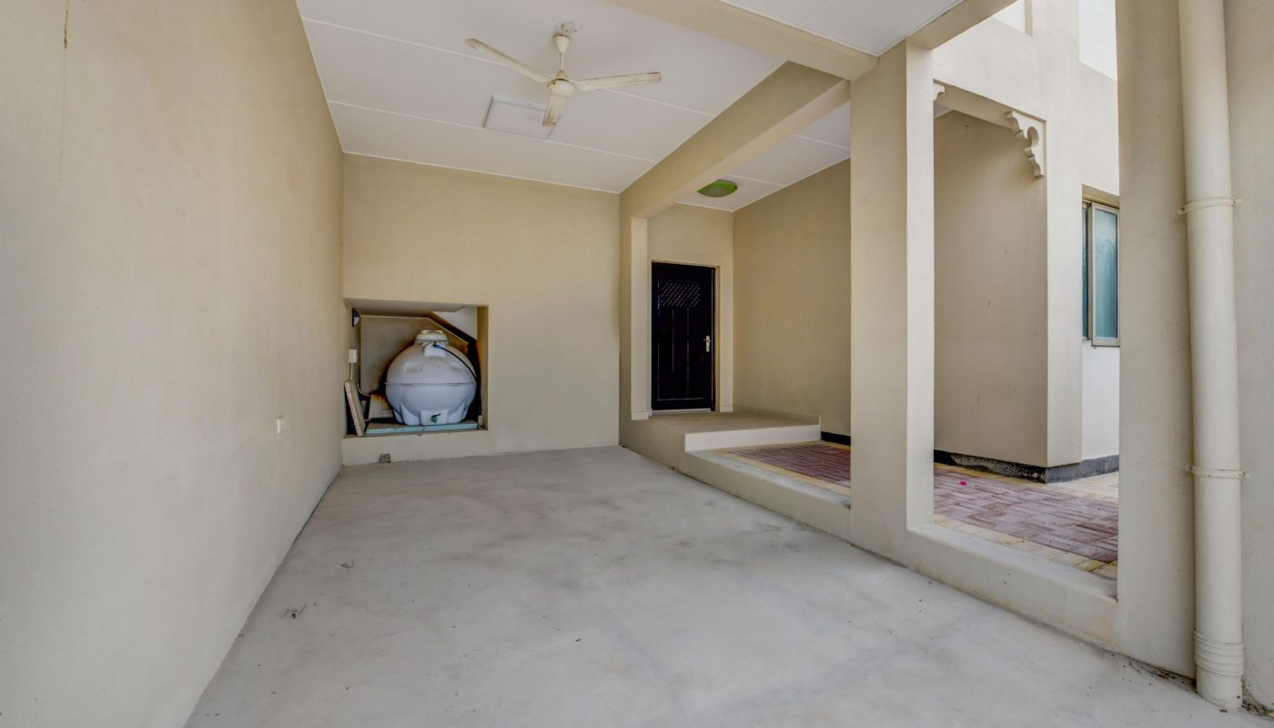 Covered area with beige walls, a ceiling fan, a storage tank in a nook, a black door, and a small outdoor tiled section with a window and white pipes.