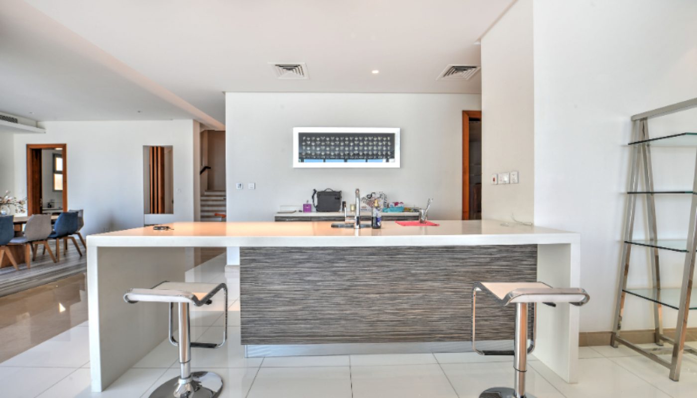 Modern kitchen with a white island, two bar stools, built-in sink, and an open view into a dining area; bright and minimally decorated space.