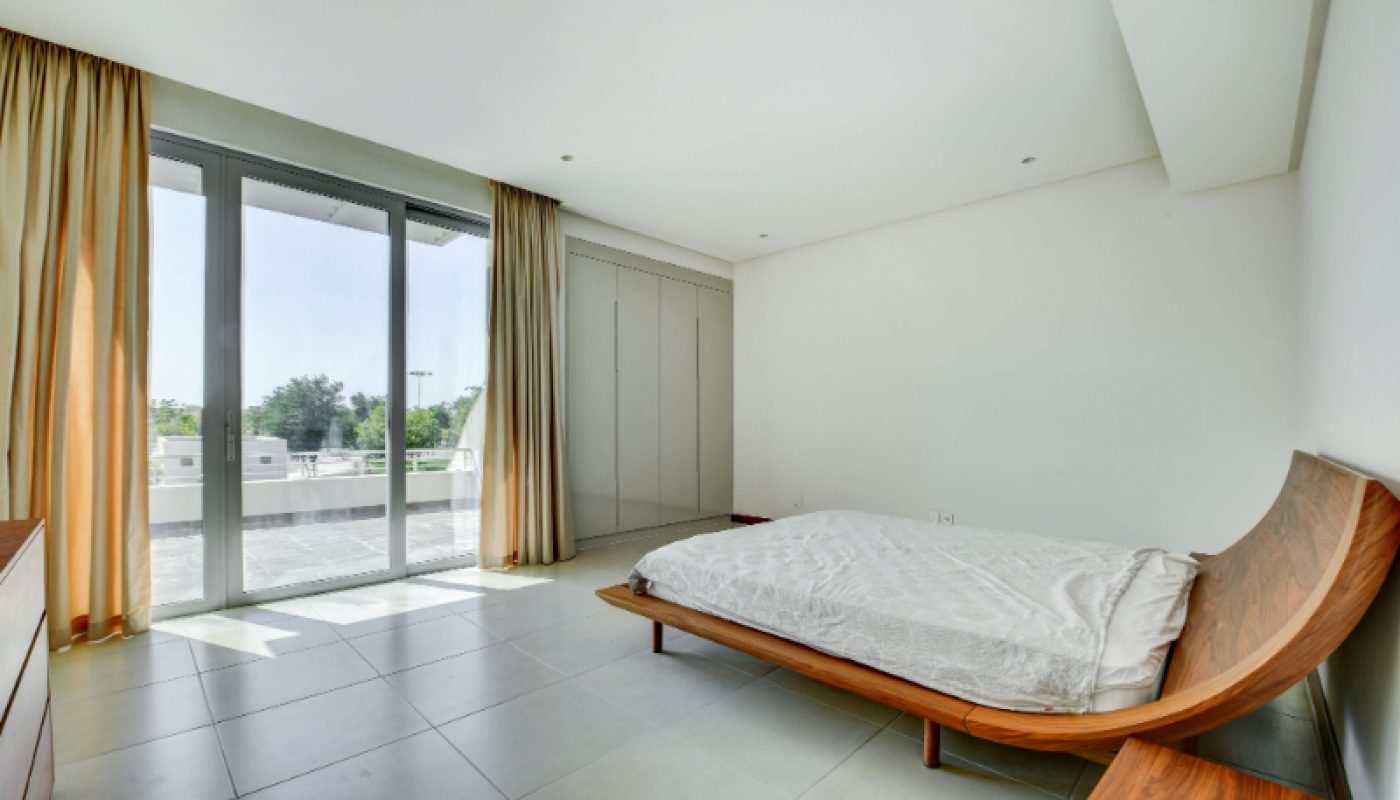 Minimalist bedroom with a wooden bed, light tile flooring, built-in wardrobe, large windows with yellow curtains, and view of a terrace outside.