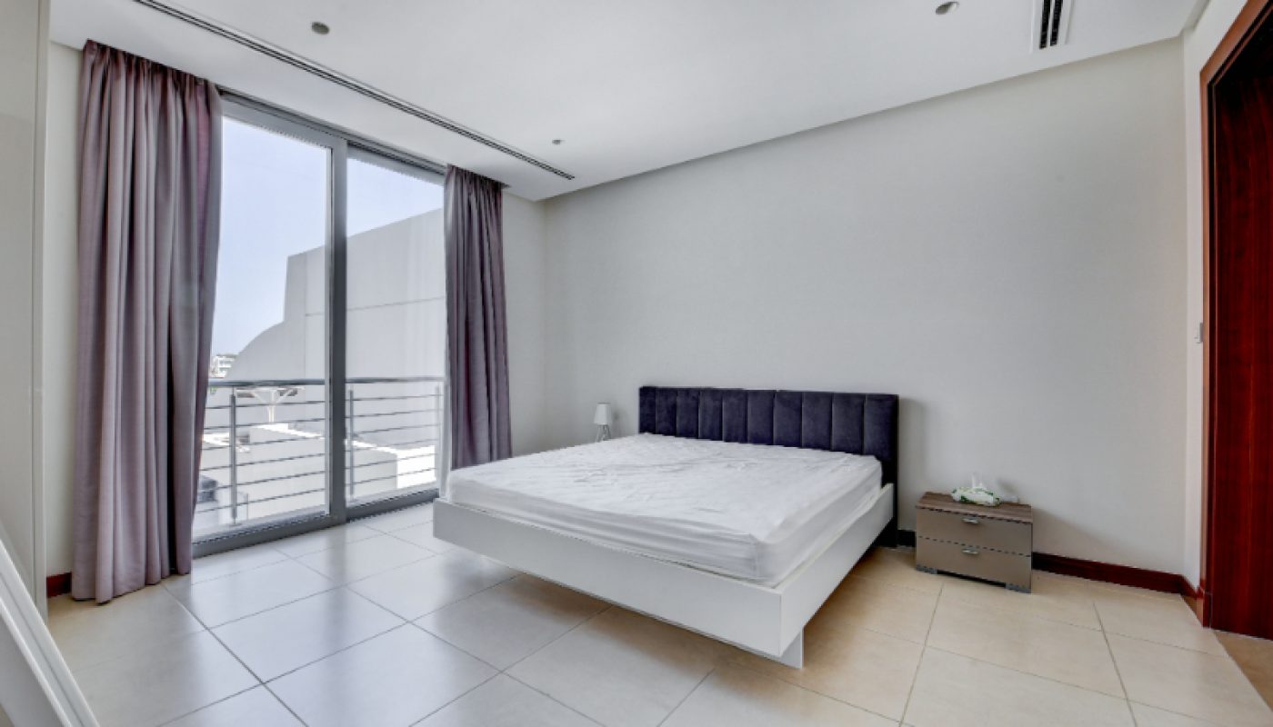 Minimalist bedroom with a large window, a bed with a dark headboard, a bedside table, neutral walls, tiled floor, and light purple curtains.