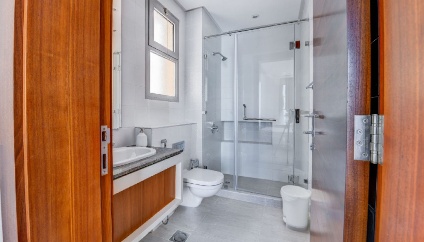 Modern bathroom with wooden door, white walls, a glass-enclosed shower, toilet, wall-mounted sink, large mirror, and small window.