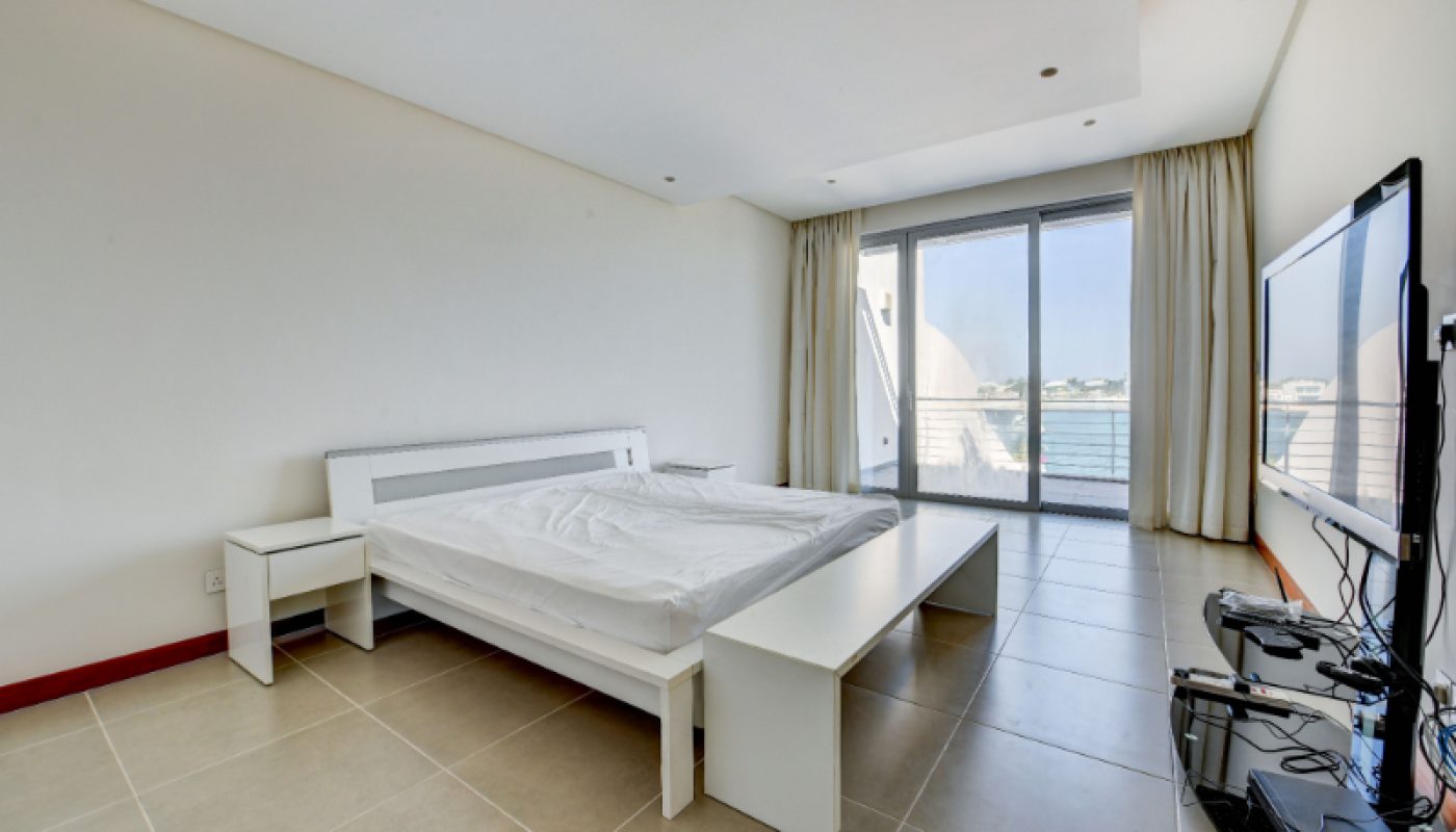Minimalist bedroom with white furniture, a large bed, a flat-screen TV, tiled floor, and a glass door opening to a balcony with a view of the sea.