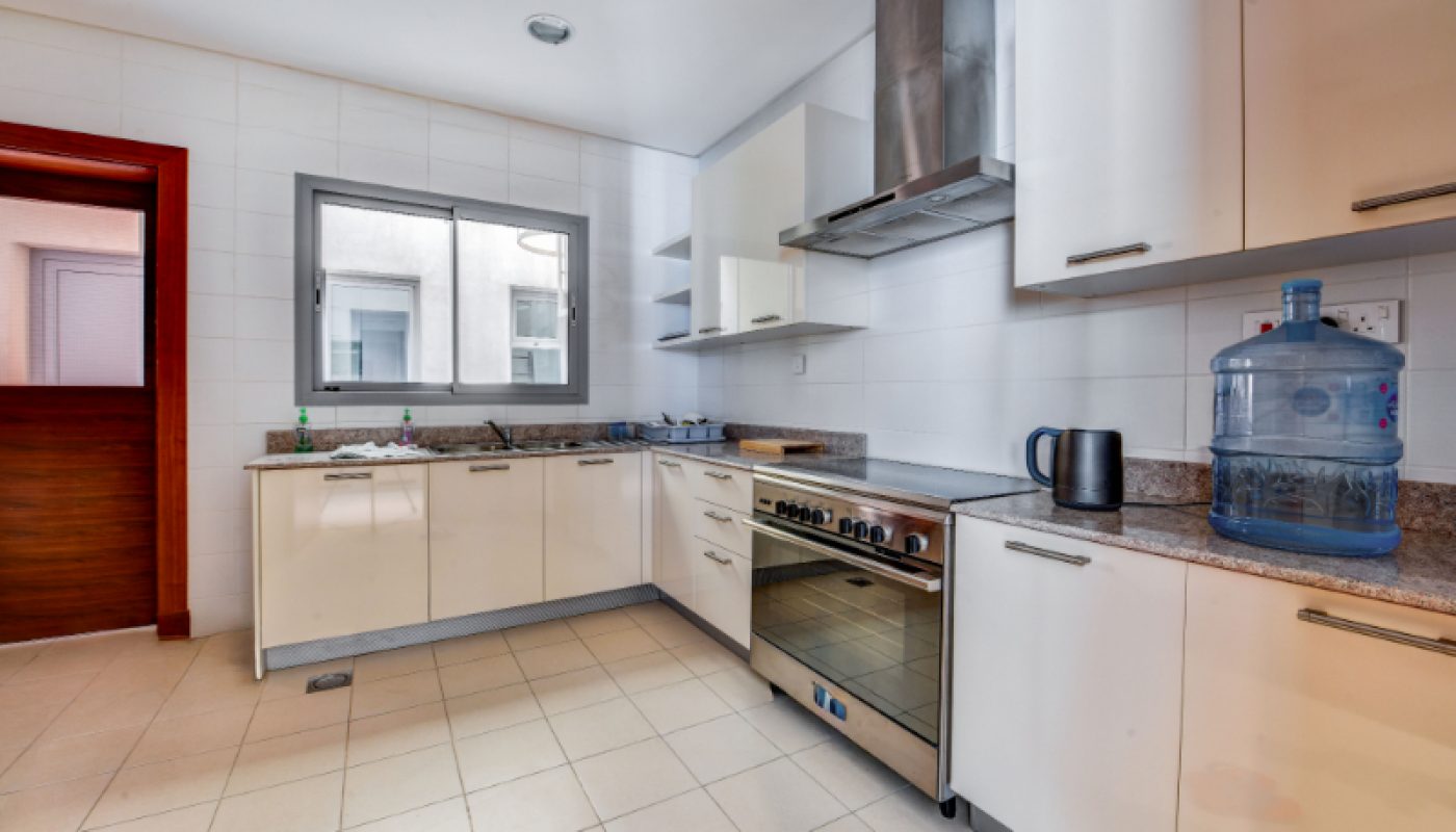 Modern kitchen with white cabinets, stainless steel oven, countertop appliances, and a large water dispenser on tiled floor; window and door visible.