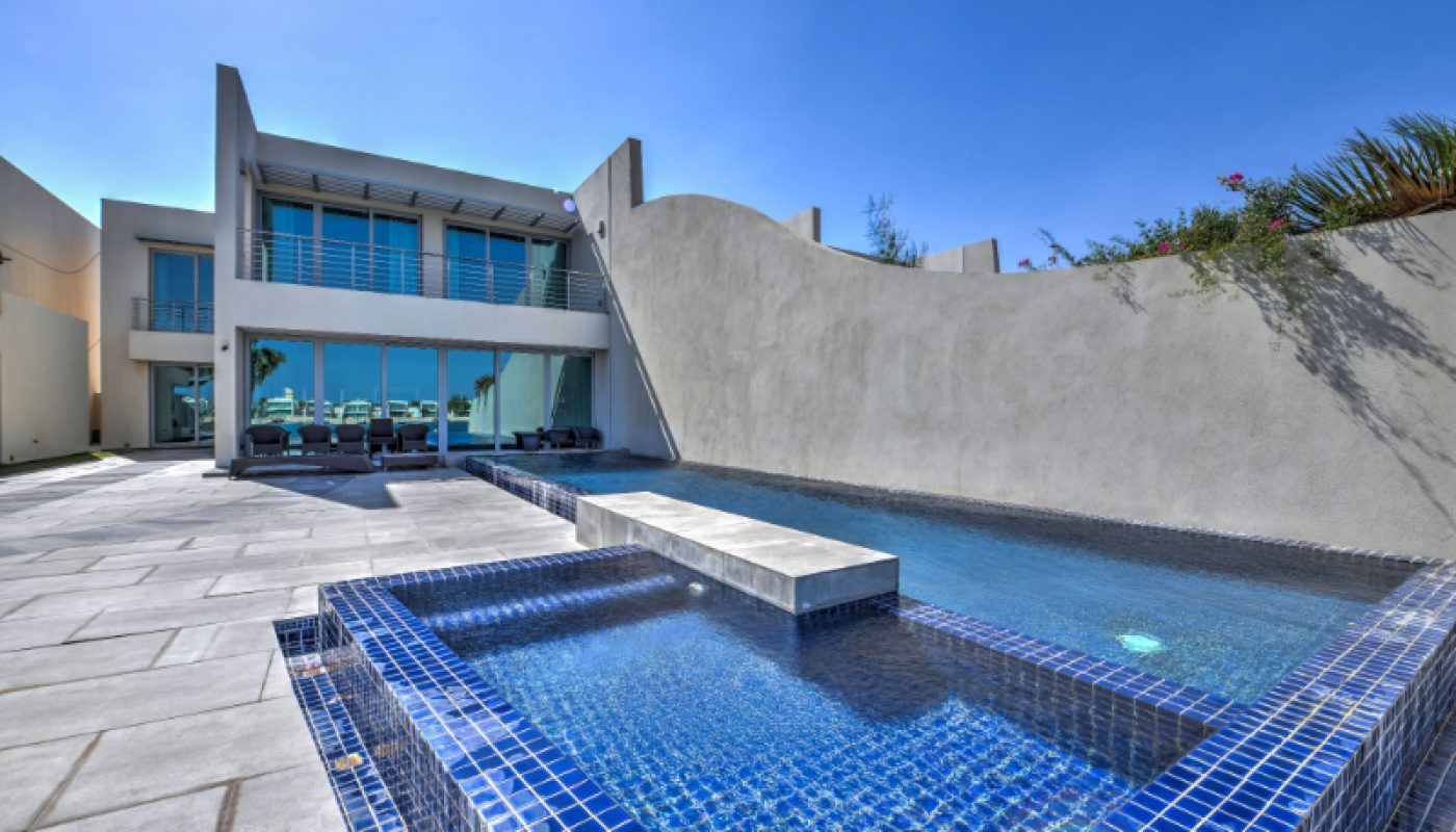 Modern house with large windows, outdoor patio, and a rectangular pool with blue tile and an attached spa under a clear blue sky.