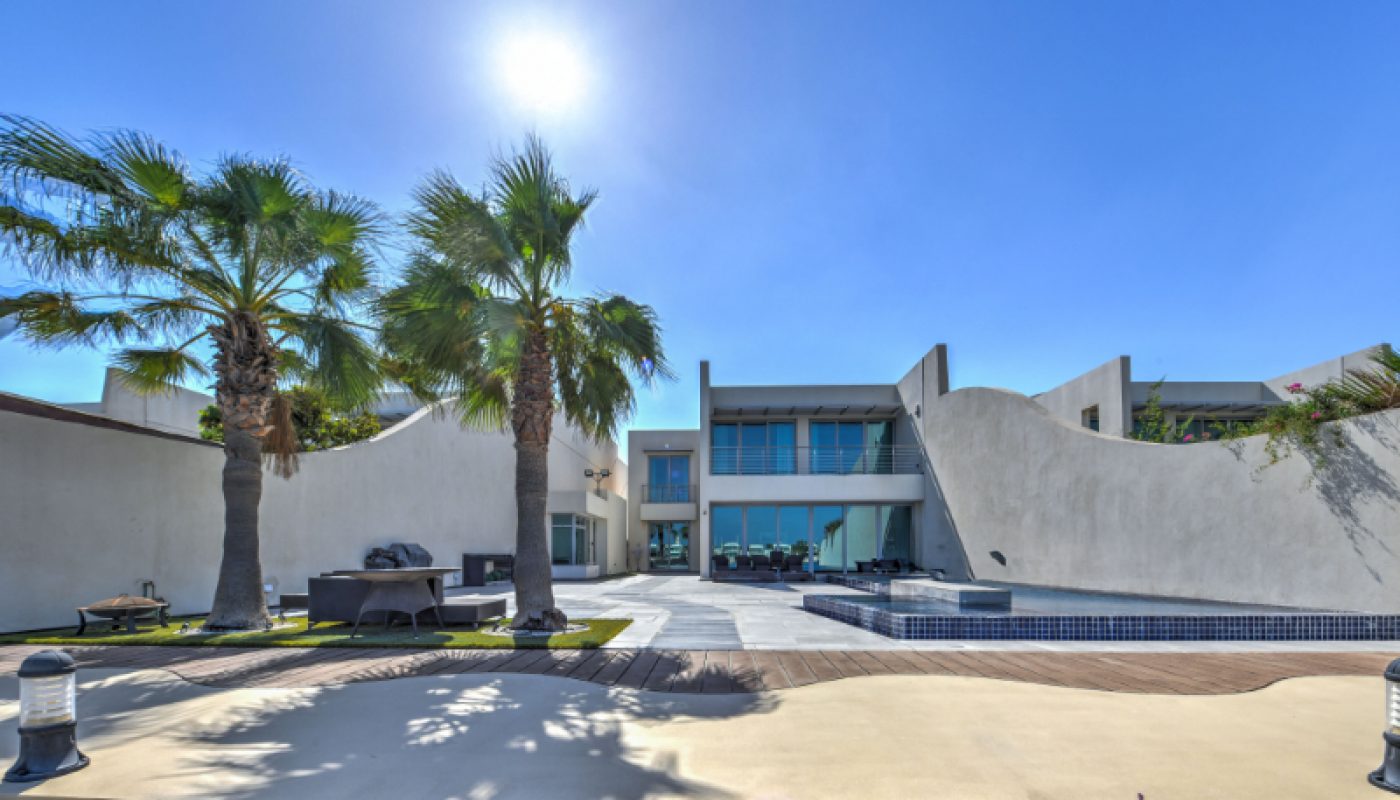 Modern two-story house with large windows, curved white exterior walls, palm trees, a patio, and an empty pool under a clear sunny sky.