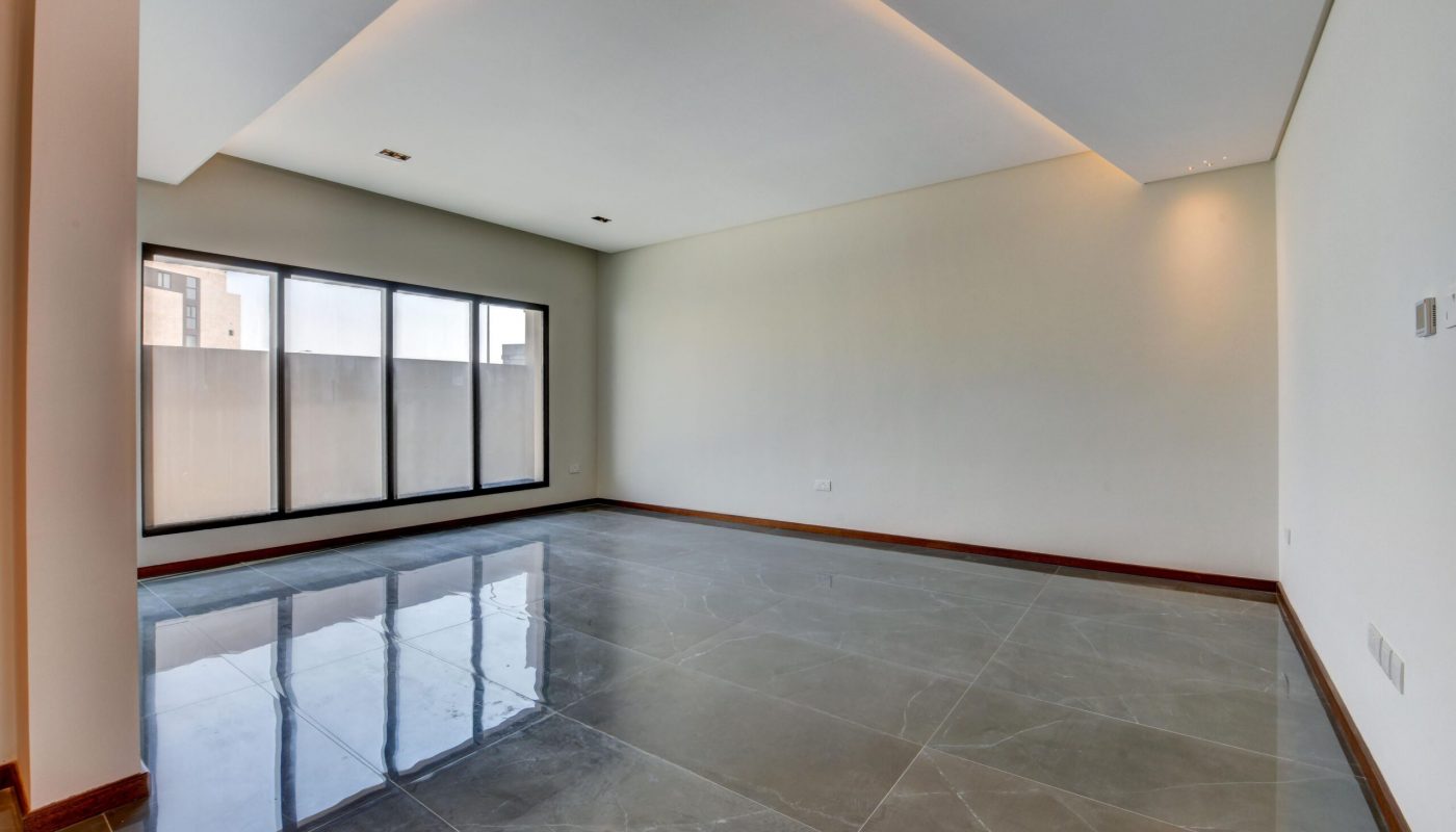 Empty modern room with large gray tile flooring, recessed ceiling lights, and floor-to-ceiling windows letting in natural light.