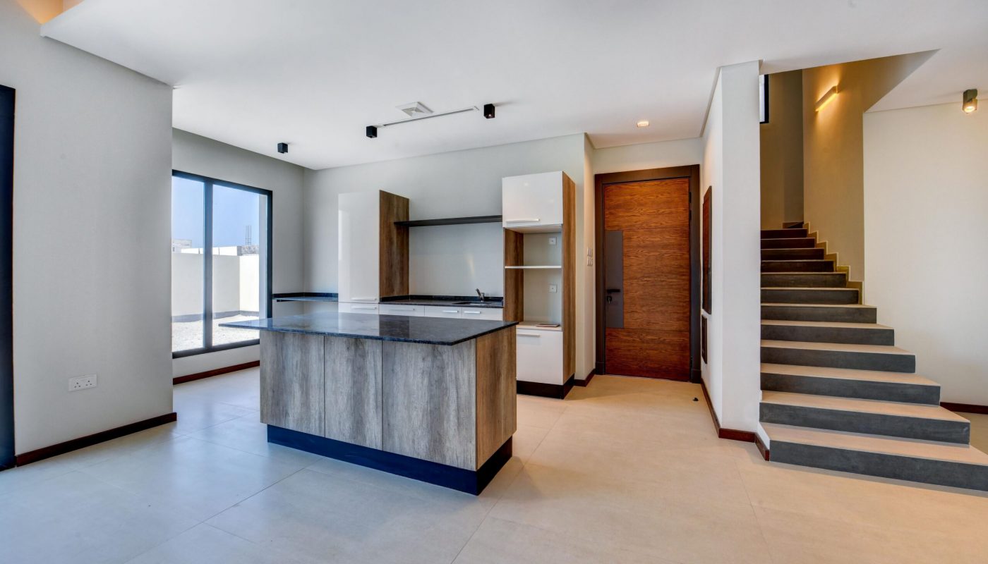 Modern kitchen with a central island, adjacent to a staircase and large windows letting in natural light. The room features neutral tones and minimalistic design.