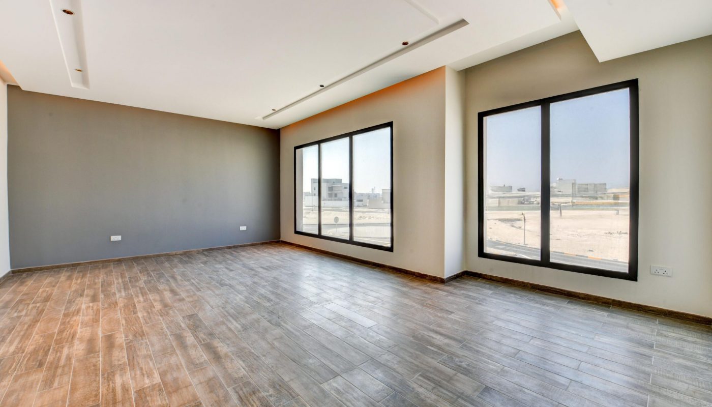 Empty modern room with large windows, gray walls, and wood-look tile flooring. Sunlight enters through the windows, showing an outdoor view of buildings and sand.