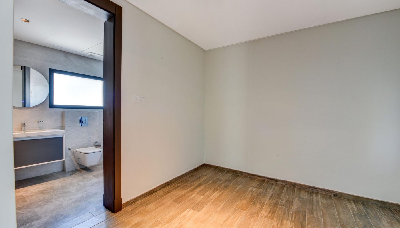 Empty room with light-colored walls and wood-look tile flooring, adjoining a bathroom with a sink, round mirror, toilet, and window.