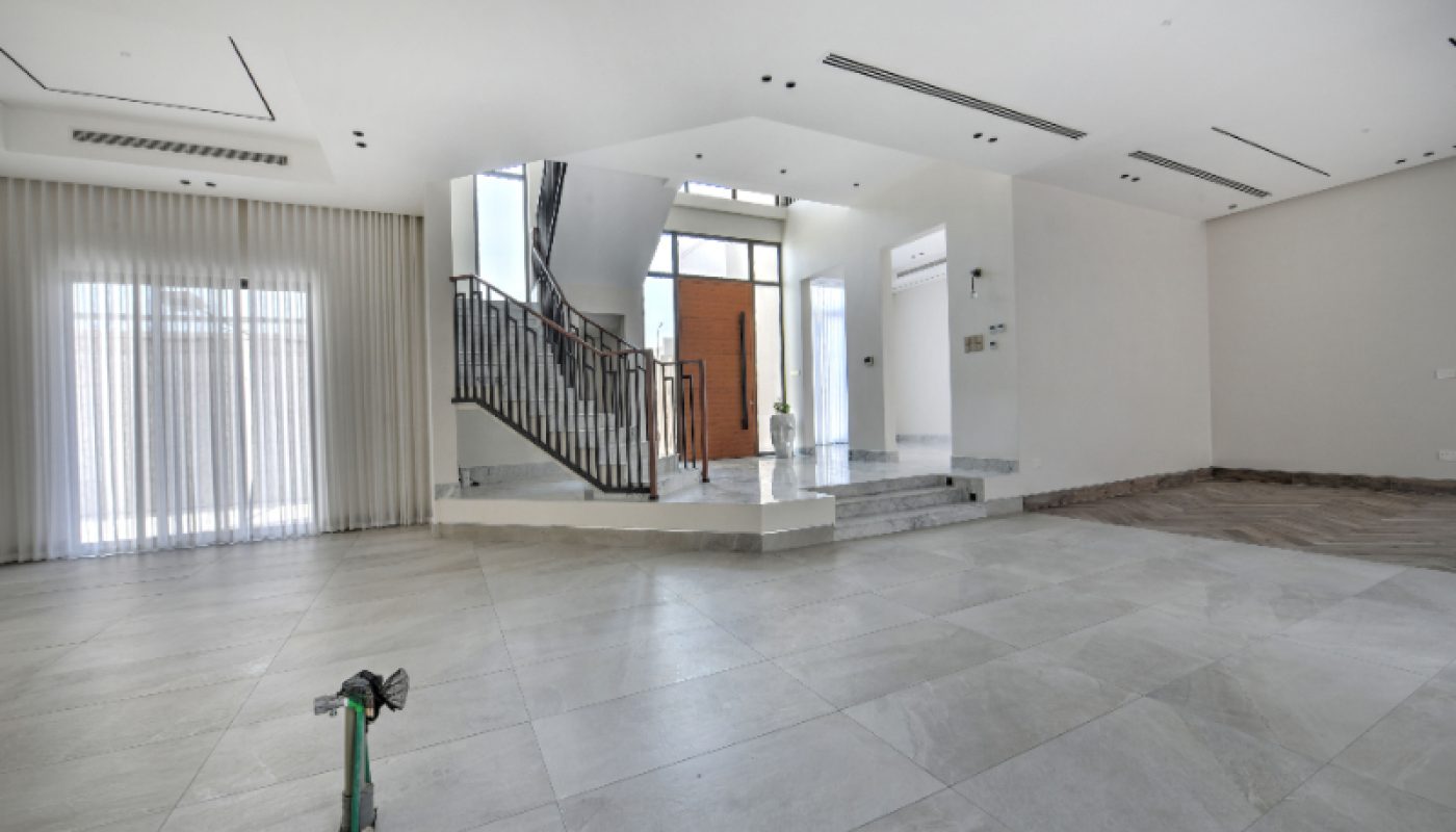 Spacious, unfurnished modern living area with large tile flooring, a staircase, floor-to-ceiling windows with sheer curtains, and pipes protruding from the floor.