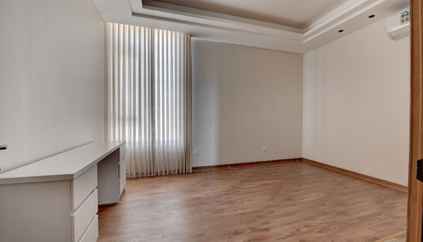 Empty room with wooden flooring, a white desk, built-in ceiling lights, large window with vertical blinds, and an air conditioning unit on the wall.