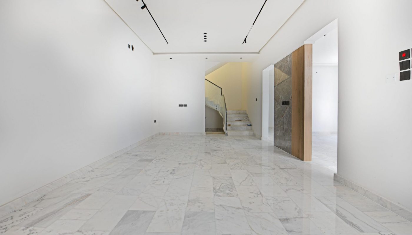Empty modern interior with white marble floors, white walls, recessed ceiling lights, and a staircase leading to an upper floor. A partially open wooden door is visible on the right.