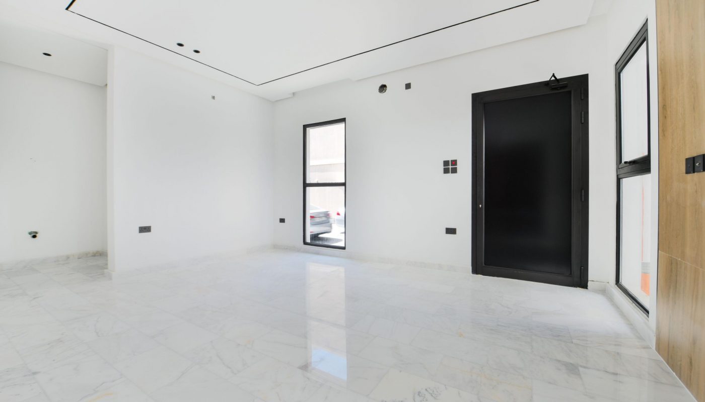 Empty modern room with white walls, marble flooring, a black door, large window, and recessed ceiling lights.