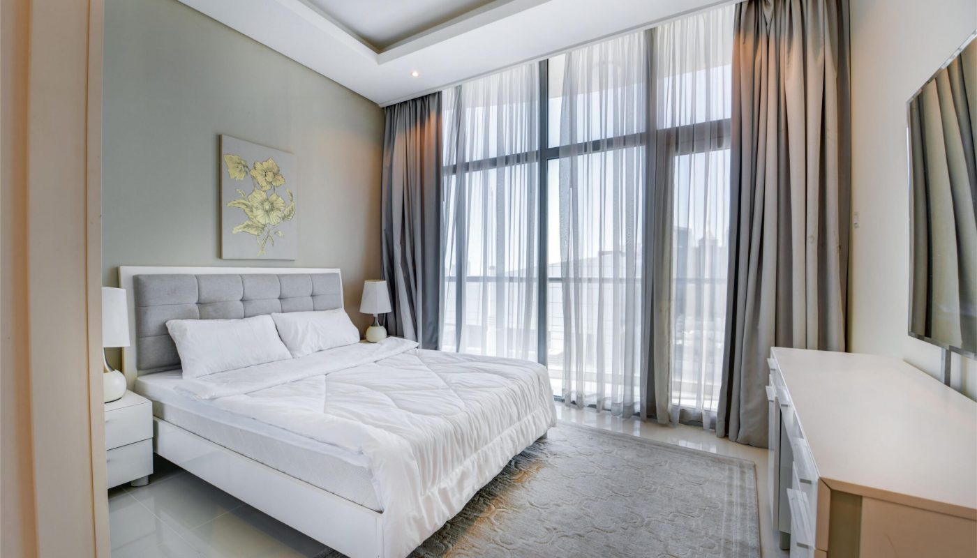 Modern bedroom with a large bed, gray headboard, two bedside tables with lamps, floor-to-ceiling windows with sheer curtains, and a wall-mounted TV.