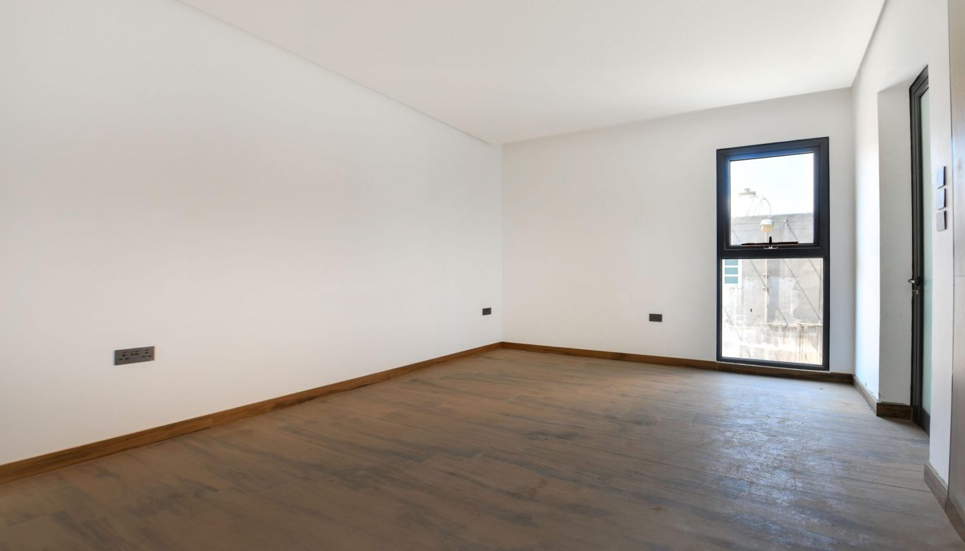 Empty room with white walls, a brown wooden floor, a single window, and a door. There are electrical outlets and natural light entering from the window.