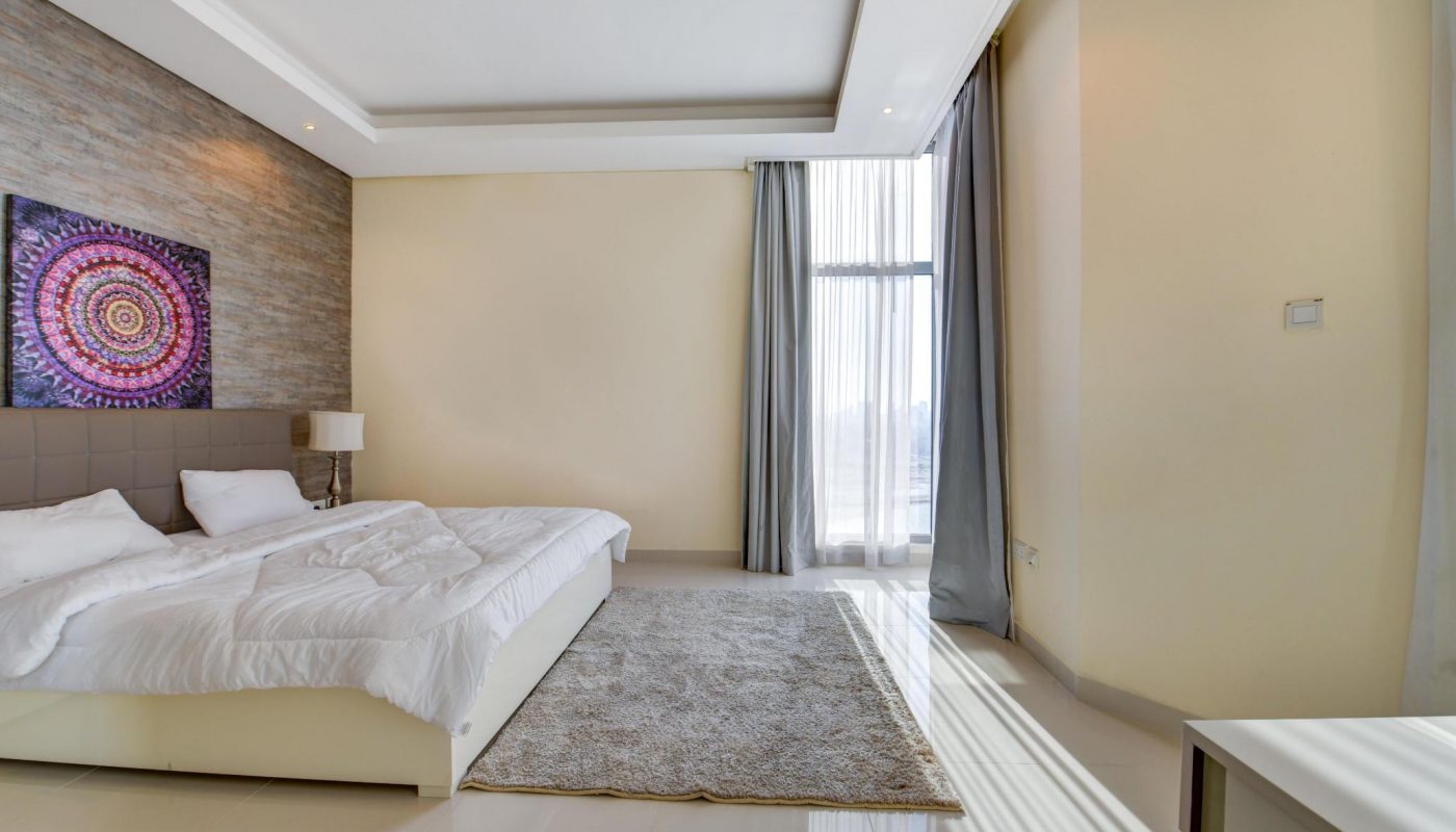 A modern bedroom with a large bed, white bedding, a nightstand with a lamp, a wall tapestry, a gray rug, and floor-to-ceiling windows with curtains letting in sunlight.