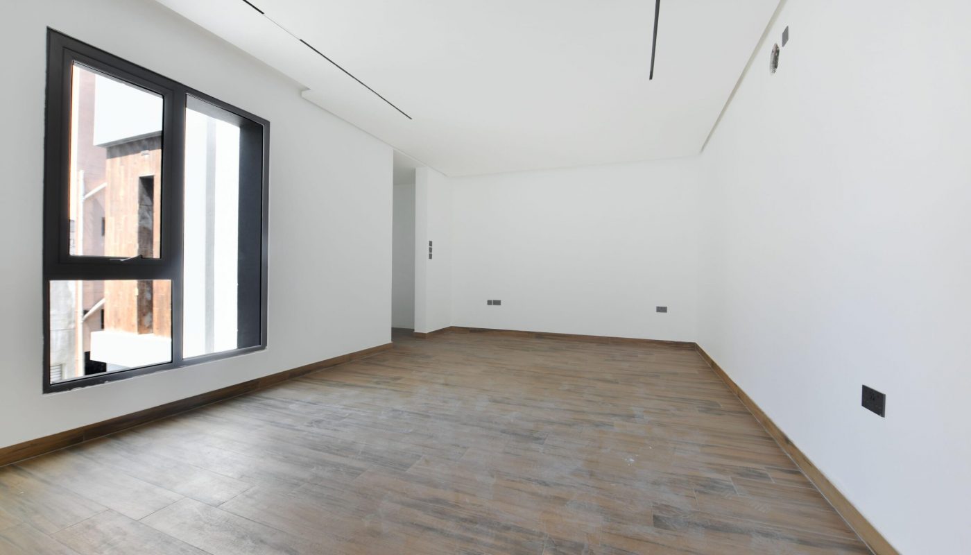 Empty room with white walls, large window, wood-patterned floor, and recessed ceiling lights.