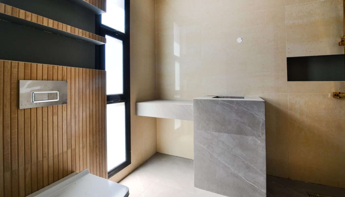 Modern bathroom with beige walls, gray stone sink, built-in shelving, and a large frosted window allowing natural light.
