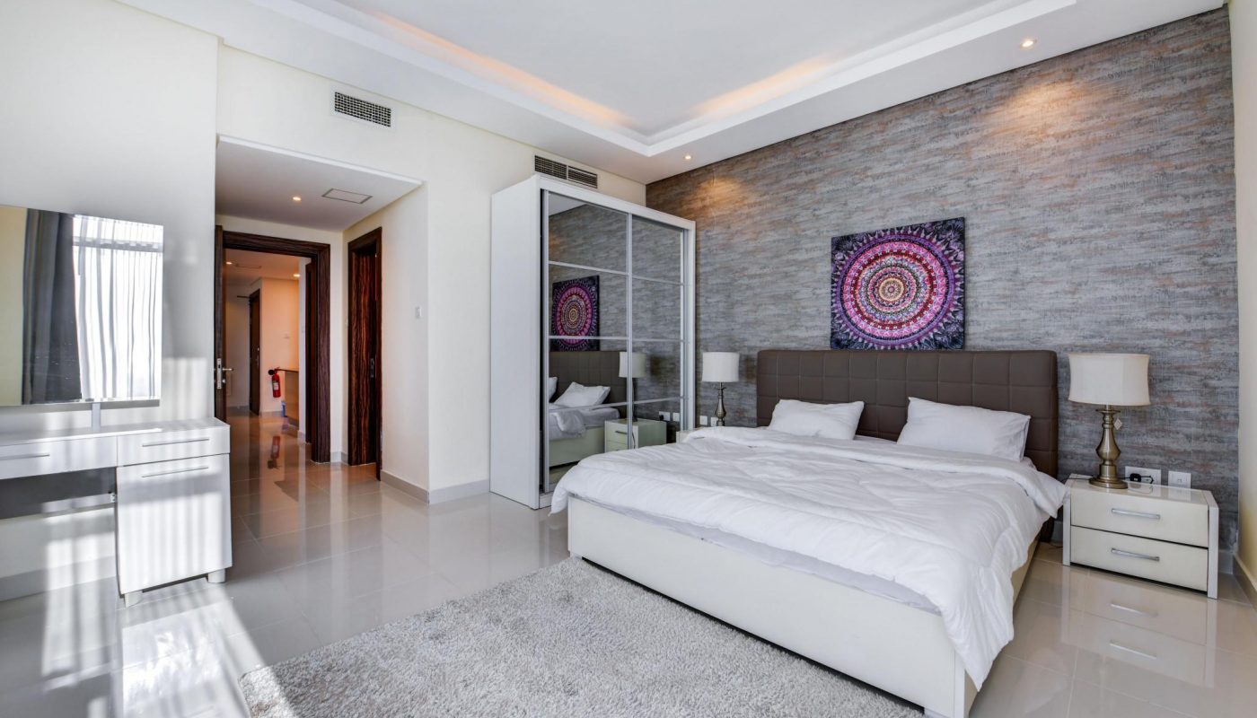 Modern bedroom with a large bed, mirrored wardrobe, textured accent wall, bedside tables, lamp, artwork above the bed, vanity, and light-colored tile flooring.