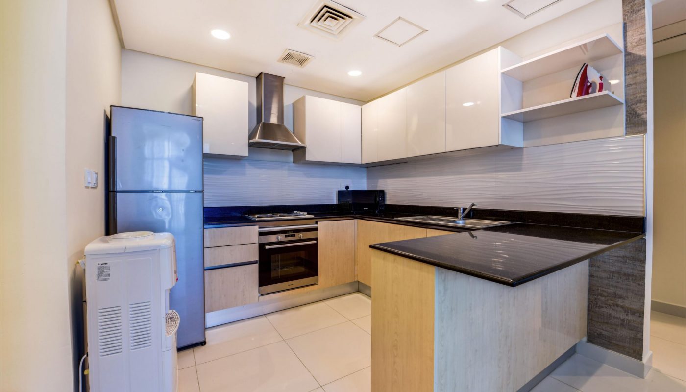 Modern kitchen with light wood and white cabinets, black countertops, stainless steel appliances, and a refrigerator; a water dispenser stands near the entrance.