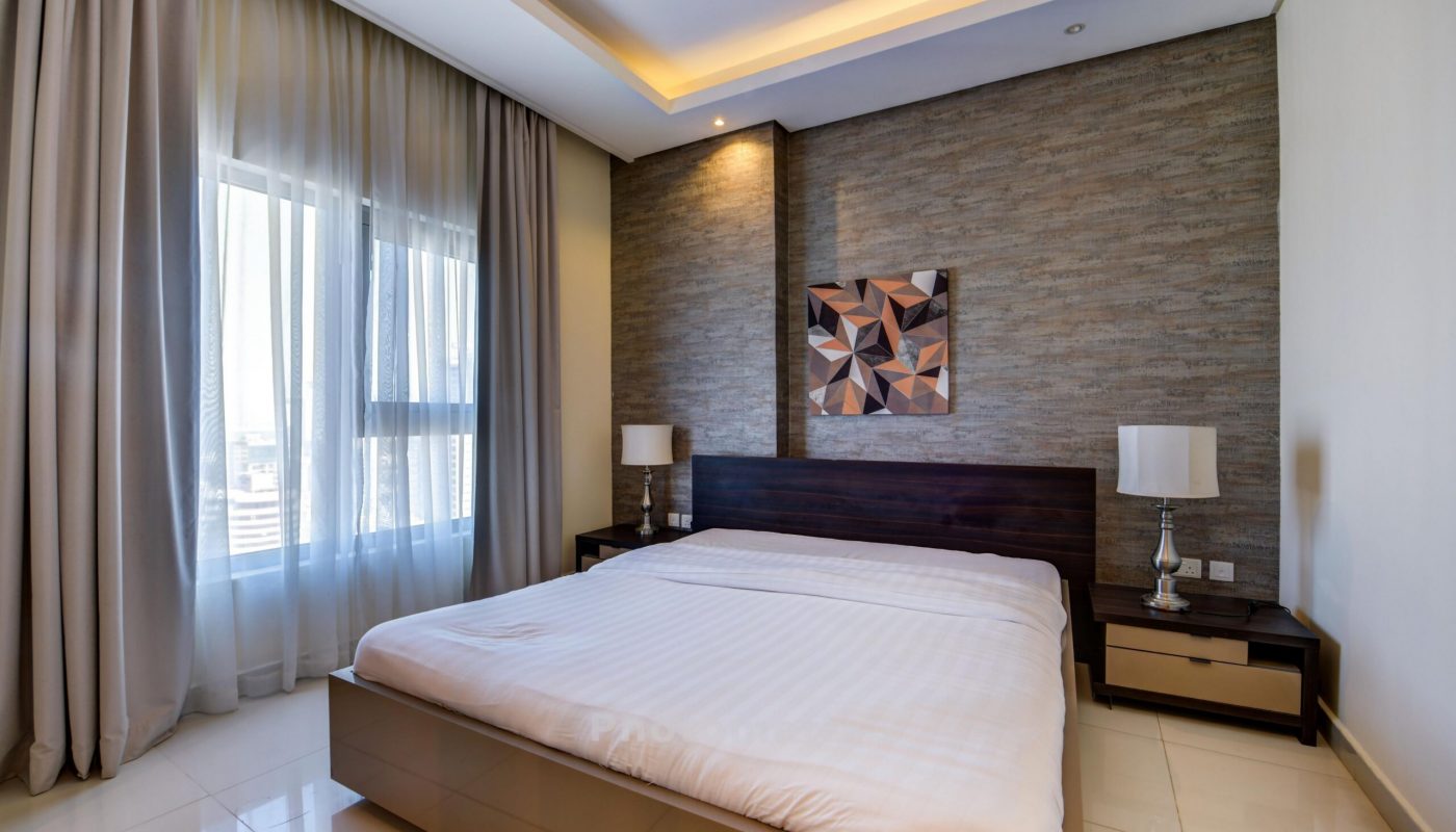 Modern bedroom with a large bed, two bedside tables with lamps, a geometric artwork above the headboard, and a window with sheer curtains letting in natural light.