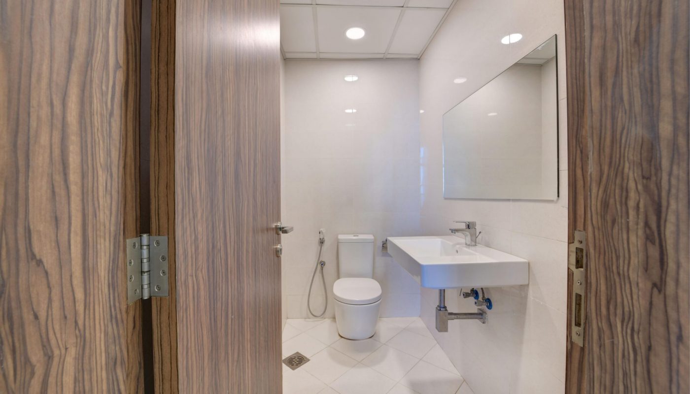 A modern bathroom with a toilet, wall-mounted sink, large mirror, bidet spray, and tiled floor, viewed through a partially open wooden door.