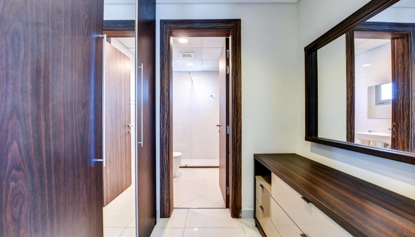 Modern bathroom with a large mirror, wooden cabinets, and an open door revealing a white-tiled shower area with a toilet.