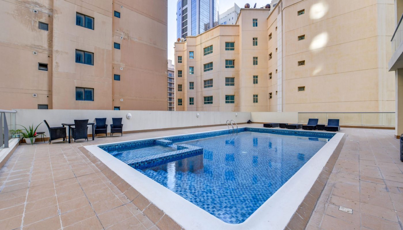 Rectangular outdoor swimming pool with blue tiles surrounded by beige buildings, poolside chairs, tables, and potted plants on tiled deck.