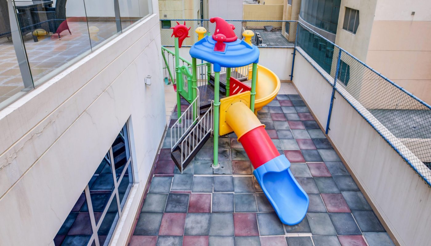 Rooftop playground with a colorful slide set on tiled flooring, surrounded by safety fencing and adjacent to building walls.