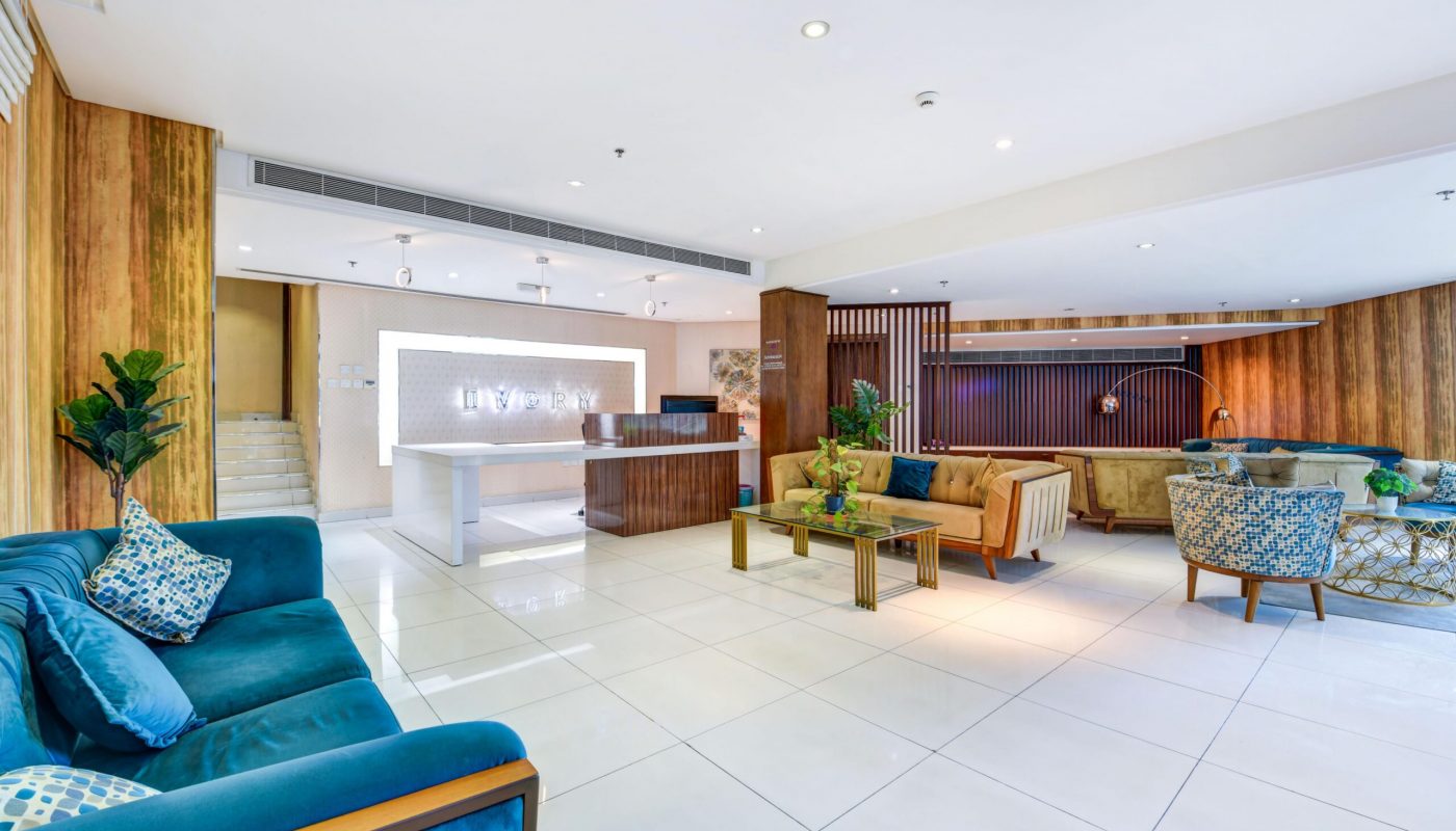 Modern hotel lobby with tiled floor, velvet sofas, coffee tables, and a reception desk with a lit sign. Indoor plants and wood panel accents decorate the space.