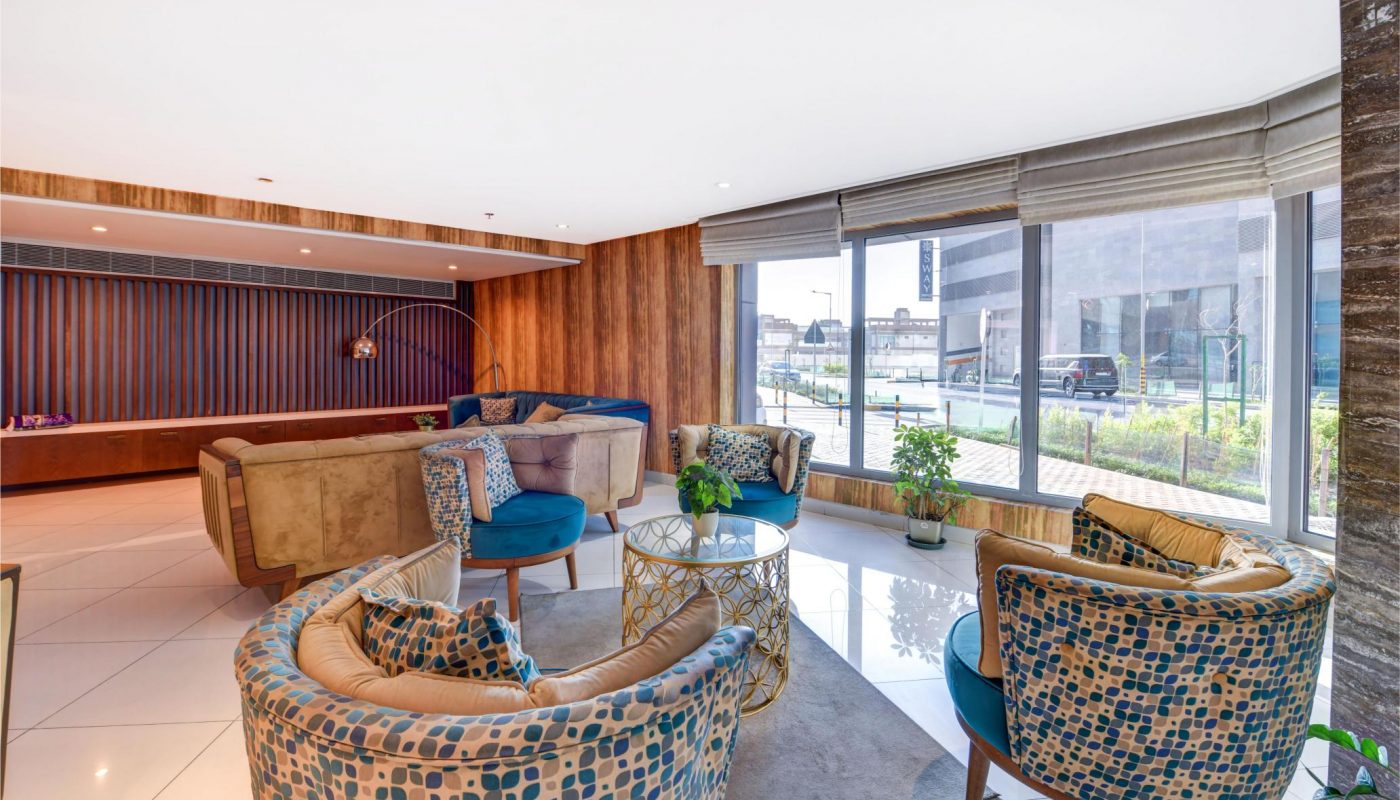 Modern lounge area with patterned armchairs, blue cushions, glass coffee table, potted plants, large windows, and wood-paneled walls.