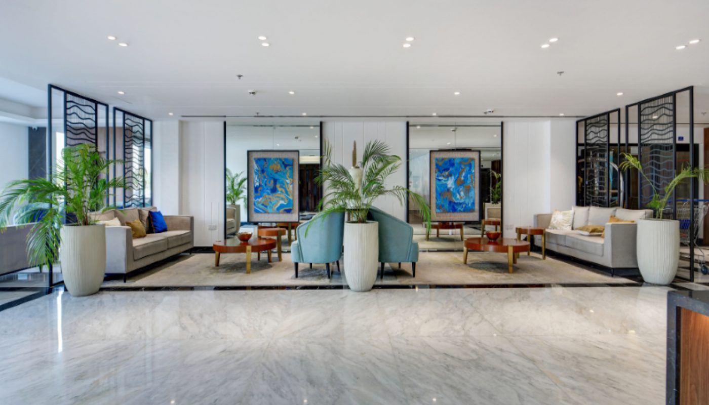 Modern hotel lobby with marble flooring, potted plants, blue chairs, sofas, and abstract paintings on the back wall.