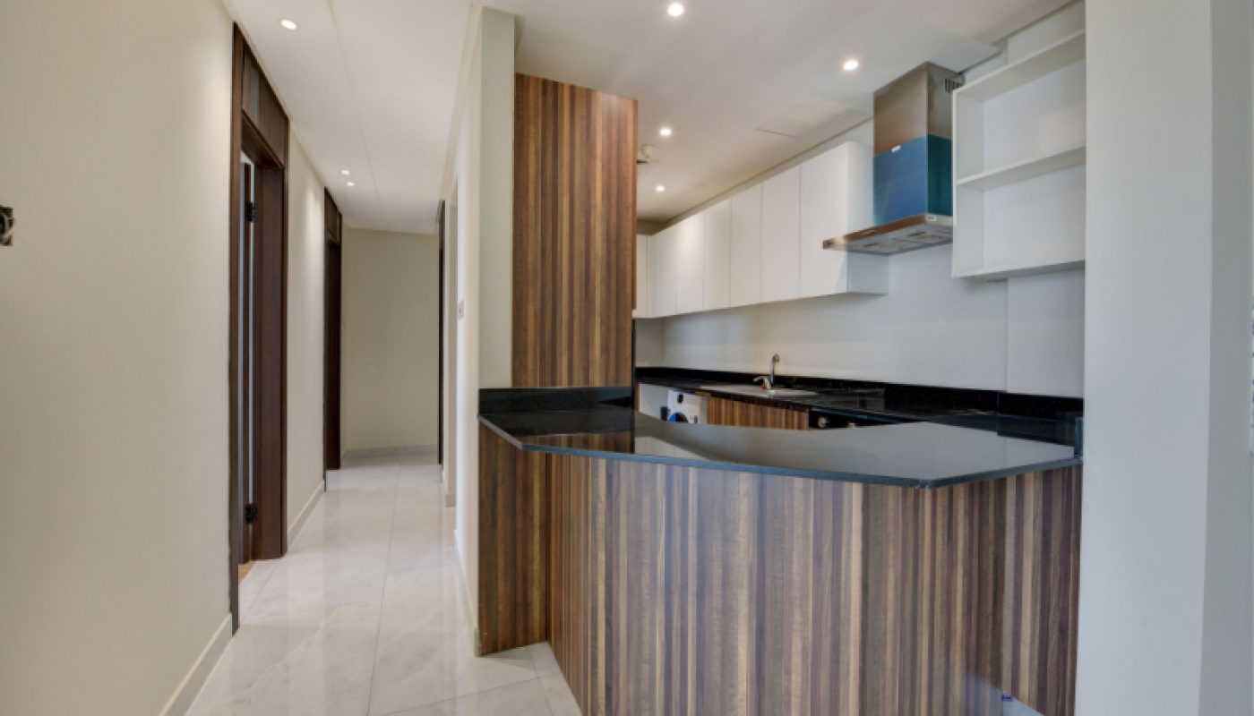 Modern kitchen with wood-paneled cabinets, black countertops, white upper cabinets, and built-in appliances, connected to a hallway with tiled flooring.