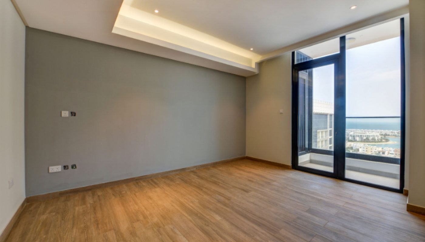 Empty modern room with wooden flooring, gray accent wall, recessed ceiling lights, and glass door leading to a balcony with a city view.