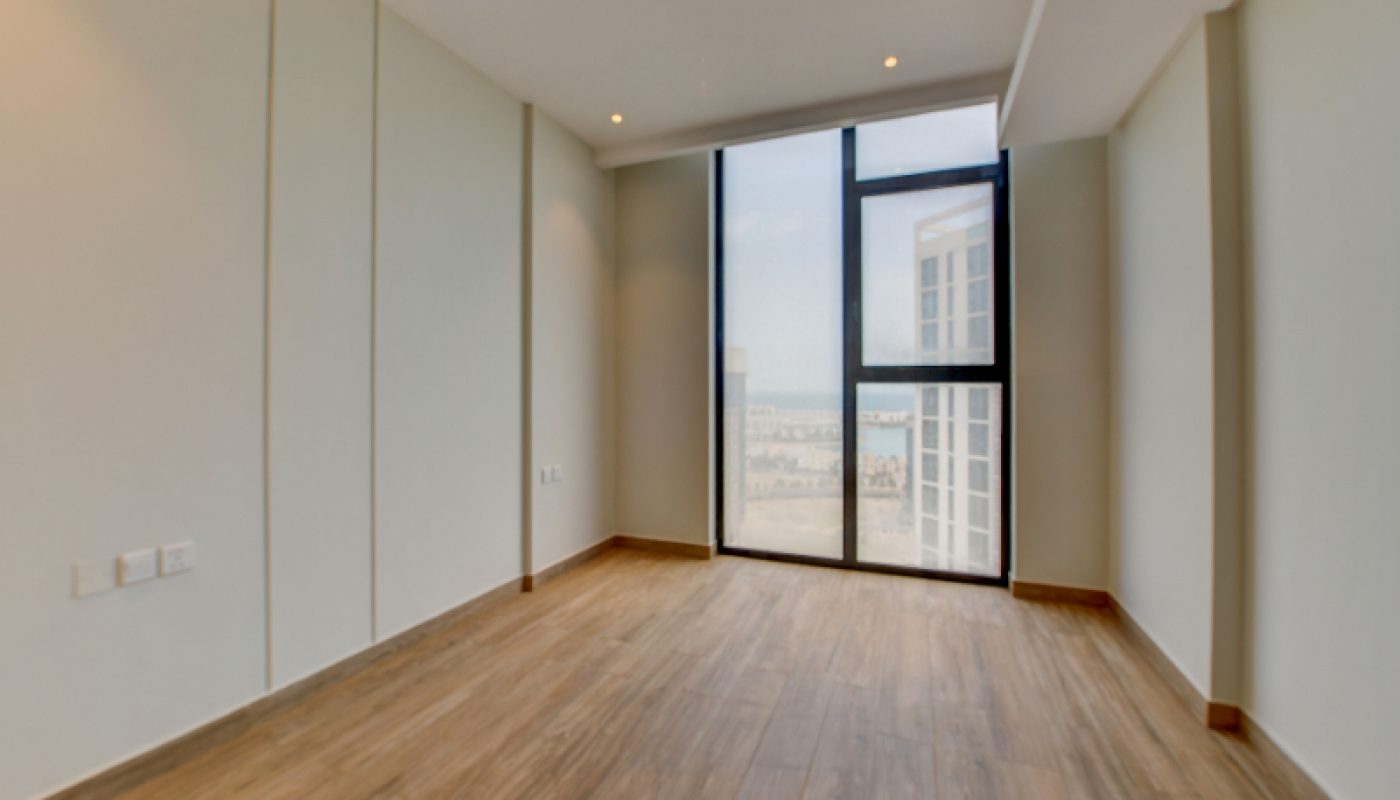 Empty room with wood flooring, cream-colored walls, recessed ceiling lights, and a large floor-to-ceiling window overlooking a cityscape and water.
